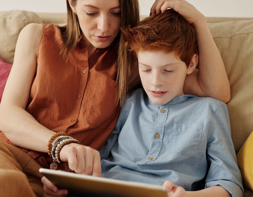 photo of woman and boy sitting on couch while using tablet computer